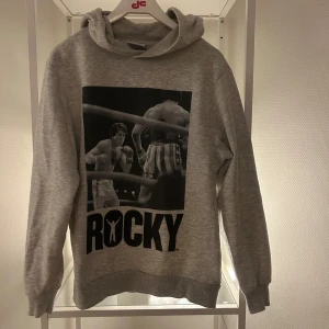 Grå Rocky hoodie från Lefties XS - Snygg grå hoodie från Lefties i storlek XS med stort Rocky-tryck både fram och bak. Tröjan har huva och ribbade muddar. Perfekt för dig som gillar filmklassiker och vill ha en bekväm streetstyle-look.