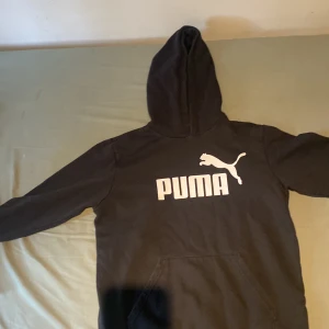 Svart hoodie från Puma med logga - Svart hoodie från Puma med stor vit logga och text på bröstet. Klassisk modell med huva och känguruficka framtill. Perfekt för en chill och sportig stil. Mjuk och skön bomullsblandning, långärmad och normal passform.