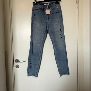 Odd Molly groupie cropped jeans - Blåa croppade jeans med broderade stjärnor, texter och coola detaljer. Råa benslut och normal passform. Jeansen har fem fickor och är tillverkade i bomull. Perfekta för dig som gillar unika och lekfulla plagg. Från märket Odd Molly i storlek W26 som ungefär motsvarar en storlek XS/S. Helt ny med tags/ prislapp fortfarande kvar. 