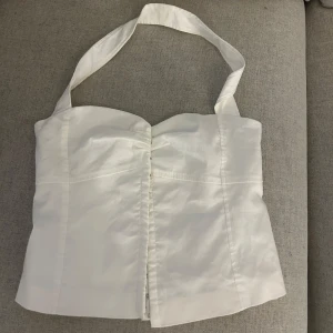 Vit korsett-topp med halterneck - Snygg vit korsett-topp från Zara med breda halterneck-band och lätt rynk framtill. Toppen har en figurnära passform och markerade sömmar som ger en stilren siluett. Perfekt att styla med jeans eller kjol. Aldrig använd.