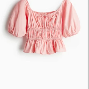 Rosa blus med puffärm och volang - Supersöt ljusrosa blus med korta puffärmar, smockad midja och volangkant nertill. Söt rosettdetalj framtill och croppad passform. Perfekt för vår och sommar, lätt och luftig i bomull.