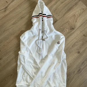 Vit hoodie från Moncler med huva - Snygg vit hoodie från Moncler med dragkedja och huva. Huvan har coola ränder i rött, blått och svart. Tröjan har långa ärmar, fickor framtill och Moncler-logga på ärmen. Perfekt för dig som gillar stilrena och sportiga plagg. Nfc scann funkar självklart. Den är i bra skick och knappt används och storlek S. 