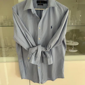 Ljusblå skjorta från Ralph Lauren - Klassisk ljusblå skjorta från Ralph Lauren med broderad marinblå logga på bröstet. Skjortan har lång ärm, knappar framtill och en snygg krage. Tillverkad i mjuk bomull och passar perfekt till jeans eller chinos. 