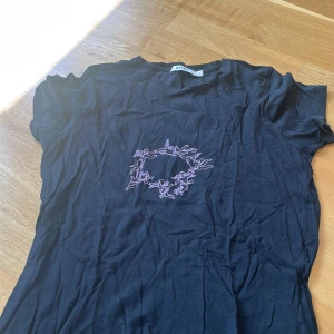 Svarta croppade toppar från NA-KD - Två svarta toppar från NA-KD i bomull. Ena toppen har ett lila broderi med älvor i cirkel på bröstet och klassisk rund hals i storlek M. Den andra är enkel, croppad och har korta ärmar. Perfekta för en chill och trendig look i storlek XS