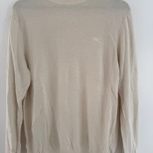 Beige stickad tröja från Zara - En stilren beige stickad tröja från Zara med rund halsringning och diskret broderad logga på bröstet. Tröjan har lång ärm och ribbade muddar vid hals, ärmslut och nederkant. 