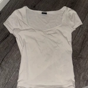 Vit topp med v-ringning från Gina Tricot - En basic vit topp från Gina Tricot med v-ringad hals och korta ärmar. T-shirten har en lätt rynkning vid bysten och är tillverkad i mjuk bomull. Perfekt att styla med jeans eller kjol för en clean look.
