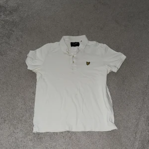 Vit pikétröja från Lyle & Scott - Snygg vit pikétröja från Lyle & Scott med klassisk krage och knäppning framtill. Tröjan har det ikoniska gula örnloggan broderad på bröstet. Perfekt för en clean och stilren look. Tillverkad i mjuk bomull för skön komfort.