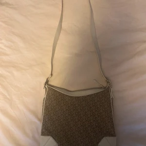 Beige och vit axelväska från DKNY - Säljer en snygg axelväska från DKNY i beige och vitt med logomönster över hela väskan. Väskan har justerbar axelrem och detaljer i vitt syntetmaterial. Perfekt för dig som vill ha en stilren accessoar med plats för det viktigaste.