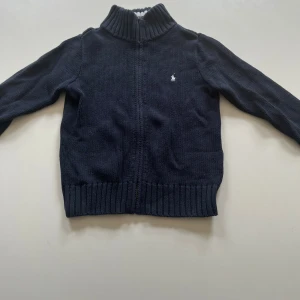 kofta från Polo Ralph Lauren - Säljer en mörkblå stickad kofta från Polo Ralph Lauren med dragkedja och vit broderad logga på bröstet. Kragen har snygga vita och blå ränder. Perfekt för kyliga dagar och enkel att matcha med det mesta. 