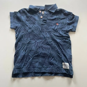 Blå piké t-shirt med mönster från H&M - Säljer en blå piké t-shirt från H&M med diskret blad-mönster och liten röd detalj på bröstet. T-shirten har krage, korta ärmar och knappar framtill. Tillverkad i 100% bomull för en skön känsla. Storlek 122/128