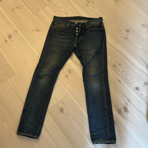 Levi's 501 jeans i mörkblå denim - Klassiska Levi's 501 jeans i mörkblå tvätt med raka ben och knäppning med knappar. Byxorna har fem fickor, tydliga slitningar och den ikoniska läderpatchen bak i midjan. Passar dig som gillar tidlös denimstil.