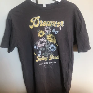 Svart t-shirt med tryck 'Dreamer' och blommor - Svart t-shirt med färgglatt tryck framtill som visar texten 'Dreamer Feeling Good' och motiv av blommor och fjärilar i gult, blått och vitt. Klassisk passform och rund halsringning. Perfekt för en avslappnad stil.