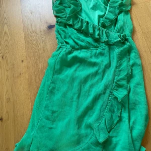 Grön omlottklänning med volang - Snygg grön omlottklänning med volangdetaljer längs urringningen och kjolen. Klänningen har en v-ringad hals och är ärmlös, perfekt för varma dagar. Materialet känns lätt och luftigt, vilket gör den bekväm att bära.