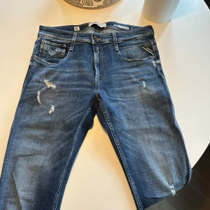 Blå Replay Anbass jeans slim fit - Säljer ett par blå Replay Anbass jeans i slim fit med klassisk femficksdesign och slitna detaljer framtill. Jeansen har normal midja och är tillverkade i ett stretchigt denimtyg för extra komfort. Perfekta för dig som gillar en smalare passform.