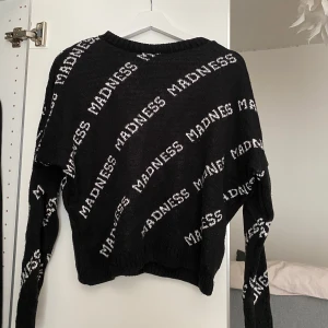 Svart stickad tröja från Madlady - Svart stickad tröja från Madlady med vit text 'MADNESS' i diagonala rader över hela tröjan. Tröjan har långa ärmar och en klassisk rund halsringning. Perfekt för dig som gillar statement-plagg och streetstyle. 