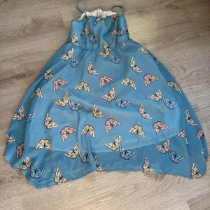 Blå klänning med fjärilsmönster från H&M - Supersöt blå klänning från H&M med tunna axelband och ett färgglatt fjärilsmönster. Klänningen har en luftig passform och är perfekt för varma dagar. Den är ärmlös och har en lätt, flowy känsla.