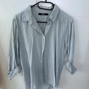 Randig skjorta från Bik Bok med broderi - Ljusblå och vit randig skjorta från Bik Bok i bomull. Klassisk krage, knäppning framtill och röd broderad text 'hello sunshine' på ryggen. Perfekt för en avslappnad stil.