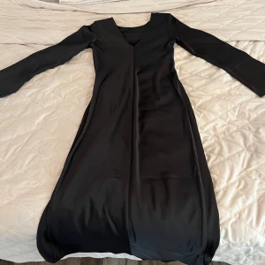 Svart långärmad klänning från H&M - Säljer en elegant svart långärmad klänning från H&M med v-ringning och snygg drapering baktill. Klänningen har en slits och en delvis figurnära passform och är tillverkad i ett glansigt, mjukt material som ger en lyxig känsla.