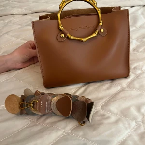 Brun handväska med bambuhandtag - Säljer en snygg brun handväska från Beverly Hills Polo Club  med unika guldfärgade bambuinspirerade handtag. Väskan har ett innerfack med dragkedja och kommer med en bred, dekorativ axelrem i olika nyanser av beige och brunt.