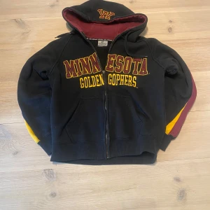 Svart Minnesota Golden Gophers hoodie - Svart hoodie med dragkedja och huva från Minnesota Golden Gophers. Broderad text i gult och vinrött framtill, vinröda detaljer på insidan av huvan och längs ärmarna samt gula inslag. Perfekt för dig som gillar college-stil. Skicka ett meddelande för frågor, storlek, mått osv. 