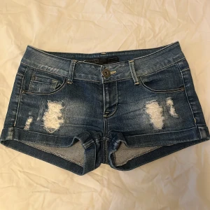 Blå jeansshorts med slitningar från Only - Snygga blå jeansshorts från Only med slitna detaljer framtill och klassisk femficksdesign. Shortsen har låg midja och korta ben, perfekt för varma dagar. Stängs med knapp och dragkedja.