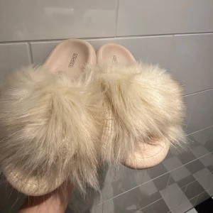 Beiga fluffiga sandaler med fuskpäls - Supermjuka och trendiga sandaler i beige med fluffig fuskpäls över foten. Perfekta för att mysa runt hemma eller för en chill look. Snygg och bekväm slip-in modell med platt sula.