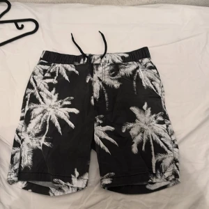 Svarta shorts med palmtryck från Kappahl - Svarta shorts från Kappahl med vitt palm-mönster. Shortsen har elastisk midja med snörning och två bakfickor. Perfekta för strand eller pool.