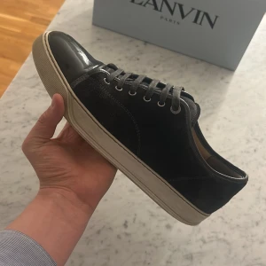 Svarta sneakers från Lanvin med lackdetaljer - Säljer ett par svarta sneakers från Lanvin med snygga lackdetaljer på tån och sidorna. Skorna har snörning och en vit platt sula. Perfekta för dig som gillar stilrena och exklusiva sneakers.