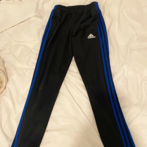 Svarta Adidas träningsbyxor med blå ränder - Svarta träningsbyxor från Adidas med klassiska blå ränder längs sidorna och broderad Adidas-logga på låret. Byxorna har resår i midjan och är tillverkade i ett mjukt och stretchigt material som passar perfekt för sport och träning. I storlek 164 13-14 år