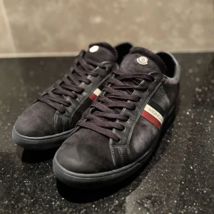 Svarta sneakers från Moncler med röd och vit detalj - Säljer ett par mörkblå/svarta Moncler sneakers med snygga röda och vita ränder på sidan samt Moncler-logga på plösen. Skorna har snörning och är tillverkade i mocka och skinn för en lyxig känsla. Perfekta för dig som gillar stilrena och exklusiva sneakers.