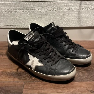 Svarta sneakers från Golden Goose med vit stjärna - Säljer ett par svarta sneakers från Golden Goose med vit stjärndetalj på sidan och vit häl. Skorna har svarta skosnören och en platt sula. Klassisk och stilren design med GGDB-logga på plösen. Nypris runt 5000 använt nån månad. Skriv om du har funderingar.