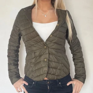 Tunn jacka - Snygg olivgrön pufferjacka med guldiga knappar framtill. Jackan har en figurnära passform och är lätt vadderad, perfekt för lite lättare kyliga dagar. Långärmad modell med enkel, stilren design som passar till många outfits.