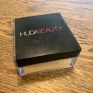 Huda Beauty Easy Bake Loose Baking & Setting Powder 'Kunafa' - Lös puder från Huda Beauty i nyansen 'Kunafa'. Perfekt för att sätta sminket och ge en matt finish. Kommer i en fyrkantig transparent burk med svart lock och tydlig märkning. Pudret är ljusgult och passar för baking och fixering.