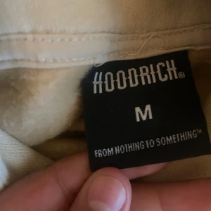 Beige och vit hoodie från Hoodrich - Säljer en beige och vit hoodie från Hoodrich med stor logga på bröstet och ärmen. Hoodien har dragkedjefickor framtill och en luva. Perfekt för en avslappnad och sportig stil.