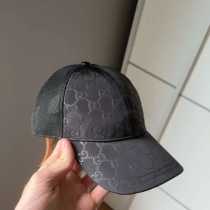 Svart keps från Gucci med GG-mönster - Snygg svart keps från Gucci med klassiskt GG-monogram över hela kepsen. Framsidan är i glansig polyester och baksidan har meshpanel för extra ventilation. Perfekt accessoar för en trendig look.