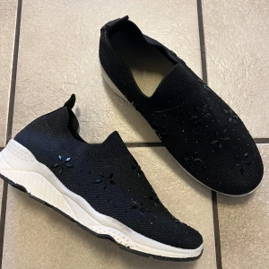 Svarta slip-on sneakers med blommiga detaljer - Säljer ett par svarta slip-on sneakers med vita sulor och blommiga dekorationer på ovandelen. Skorna är stickade och har en sportig, bekväm design utan snörning. Perfekta för dig som gillar stilrena och unika detaljer.