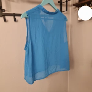 Blå ärmlös blus - Säljer en blå ärmlös blus i tunn och luftig polyester. Blusen har v-ringning och är croppad, perfekt för varma dagar. Enkel och stilren design som passar till mycket.