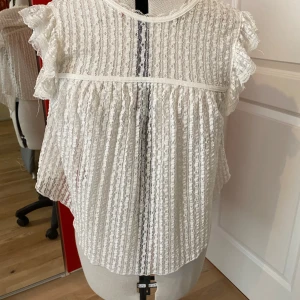 Vit spets blus från Nellie - Säljer en söt vit blus i spets med volangdetaljer på ärmarna och knytning i nacken. Blusen har korta ärmar och ett romantiskt uttryck med genombrutet spetsmönster. Perfekt för dig som gillar feminina detaljer! Endast använd fåtal gånger