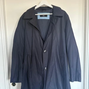 Mörkblå rock från Hugo Boss - Säljer en stilren mörkblå rock från Hugo Boss med klassisk krage och knäppning framtill. Jackan är vattentät och har två snedställda fickor och har ljusblå detaljer på insidan. Perfekt för dig som vill ha en tidlös och elegant look. 