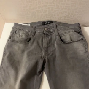 Grå Replay Anbass jeans - Säljer ett par grå Replay Anbass jeans i slim fit-modell. Byxorna har klassisk femficksdesign, dragkedjegylf och snygga slitningar för en cool look. Perfekta för dig som gillar stilrena och bekväma jeans.