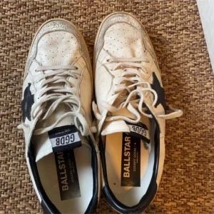 Vita sneakers från Golden Goose Ballstar - Säljer ett par vita Golden Goose Ballstar sneakers med svart stjärndetalj på sidan och grå snörning. Skorna har perforerad tå och beige sula. Ikonisk vintage-look och bekväm passform. Kommer med originalpåse.
