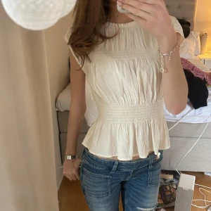 Krämvit blus med volang - Säljer en söt krämvit blus med smockad midja och volang nertill. Blusen har korta ärmar och rund halsringning. Perfekt till jeans eller kjol! 🌷🥰💕