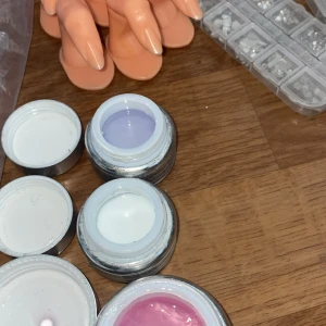 Nagelgel och tillbehör för nageldesign - Flera burkar nagelgel i vitt, ljuslila, ljusblått och rosa, tillsammans med plasttips, konstgjorda övningsfingrar och askar med nagelstenar. Perfekt för att skapa kreativa nageldesigner hemma. Nagelkudde medföljer Allt är från Nagelgiganten