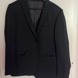 Svart kostym från Dressmann Slim Fit - Säljer en svart kostym från Dressmann i slim fit-modell, storlek 46 regular. Kavajen har klassisk design med knappar och byxorna har diskreta detaljer med röd insida vid linningen. Perfekt för formella tillfällen.