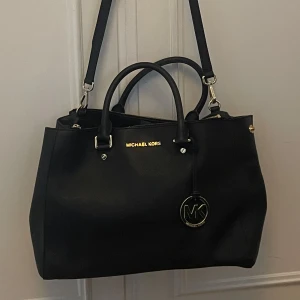 Svart handväska från Michael Kors - Snygg svart handväska från Michael Kors med både axelrem och handtag. Väskan har guldfärgade detaljer och en rund MK-berlock framtill. Perfekt för att bära med sig det viktigaste och lite till.