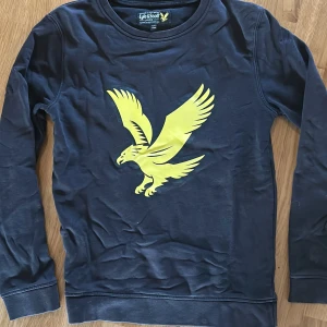 Mörkblå långärmad tröja från Lyle & Scott - Säljer en mörkblå långärmad tröja från Lyle & Scott Junior med en stor gul örn på bröstet. Tröjan har rund halsringning och är tillverkad i mjuk bomull. 