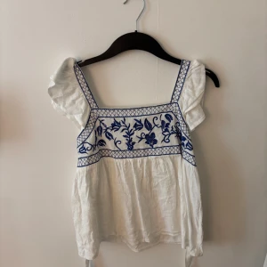 Vit blus med blå broderi och volangärmar - Superfin vit blus med blått broderat blommönster över bysten och volangärmar. Blusen har fyrkantig ringning och är tillverkad i ett lätt material. Man knyter den i ryggen, super söt😊