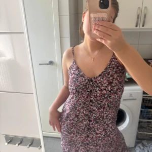Blommig sommarklänning med smala axelband - Säljer en söt blommig klänning med smala axelband och v-ringning. Klänningen har en svart bas med små rosa, vita och gröna blommor. Perfekt för varma dagar och har en lätt, luftig känsla.