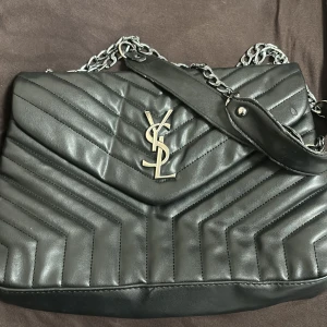 Svart axelväska från Yves Saint Laurent - Säljer en svart axelväska från Yves Saint Laurent med quiltad design och silverfärgad kedja. Väskan har YSL-logga i metall framtill och är tillverkad i skinn. Perfekt för dig som vill ha en stilren och lyxig accessoar.  !!EJ ÄKTA!!