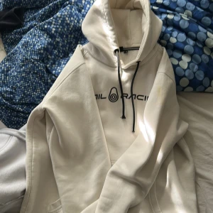 Vit hoodie från Sail Racing - Säljer en vit hoodie från Sail Racing med svart tryck på bröstet och svarta snören i huvan. Tröjan har känguruficka och en avslappnad passform, perfekt för en chill stil. Finns en gul fläck som syns på bilden som förmodligen går bort i tvätten.